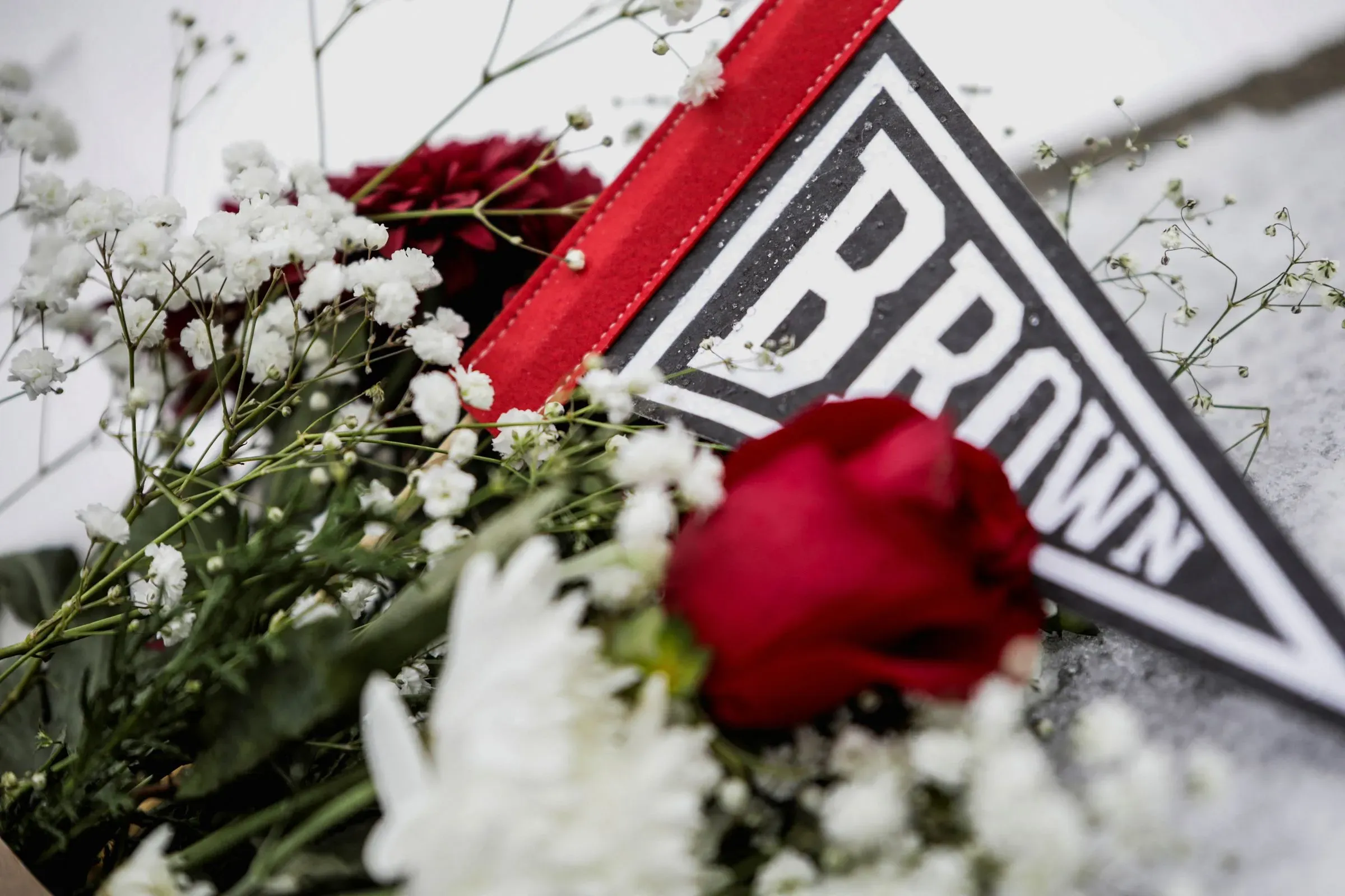 تجمع زهور حمراء وبيضاء مع علم يحمل اسم "براون"، مما يعكس الحزن والقلق في مجتمع الجامعة بعد حادث إطلاق النار.