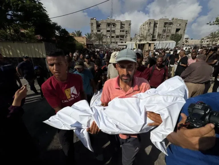 مواطنون فلسطينيون يحملون جثمانًا ملفوفًا في قماش أبيض وسط حشود في غزة، في سياق الحرب المستمرة وتأثيرها على المدنيين.