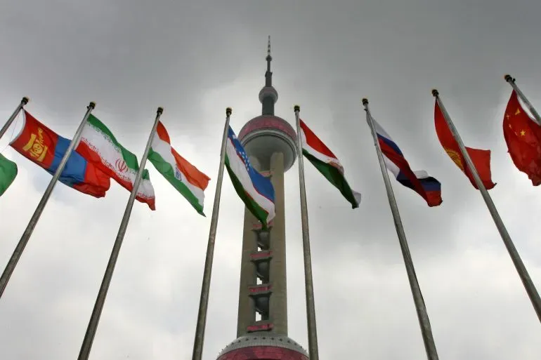 أعلام الدول الأعضاء في منظمة شنغهاي للتعاون ترفرف أمام برج في مدينة صينية، مع سماء غائمة، تعكس أهمية القمة المقبلة.