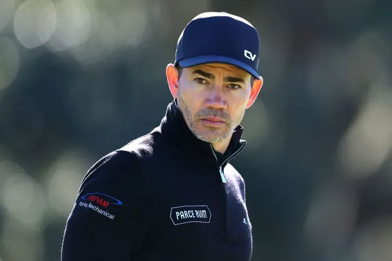 رجل يرتدي قبعة وقميص أسود، يبدو عليه التركيز والجدية، في إطار من ملعب جولف، مما يعكس تحديات رياضة الجولف.