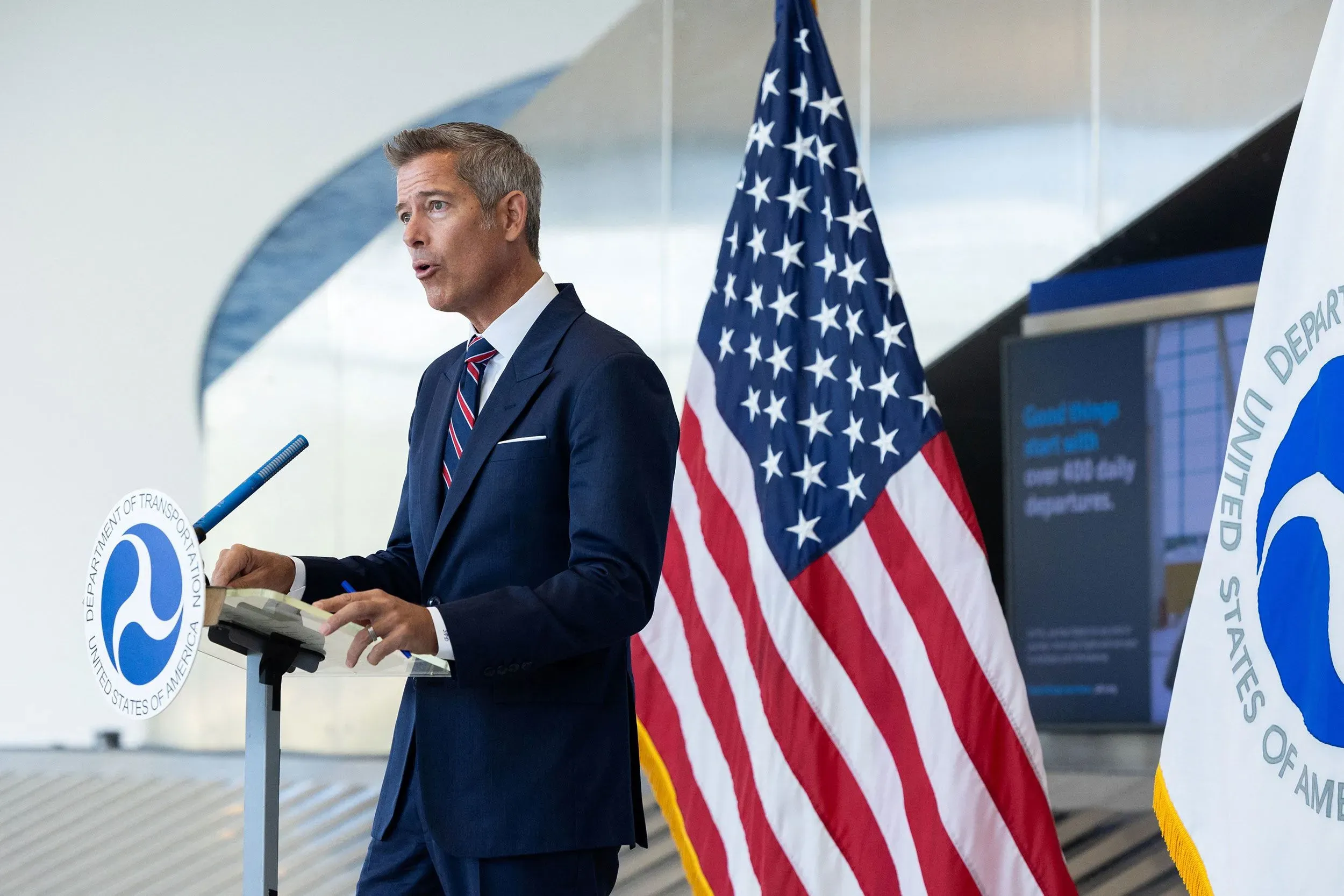 وزير النقل شون دافي يتحدث أمام ميكروفون في مؤتمر صحفي، مع العلم الأمريكي وعلم وزارة النقل خلفه، وسط أجواء رسمية.