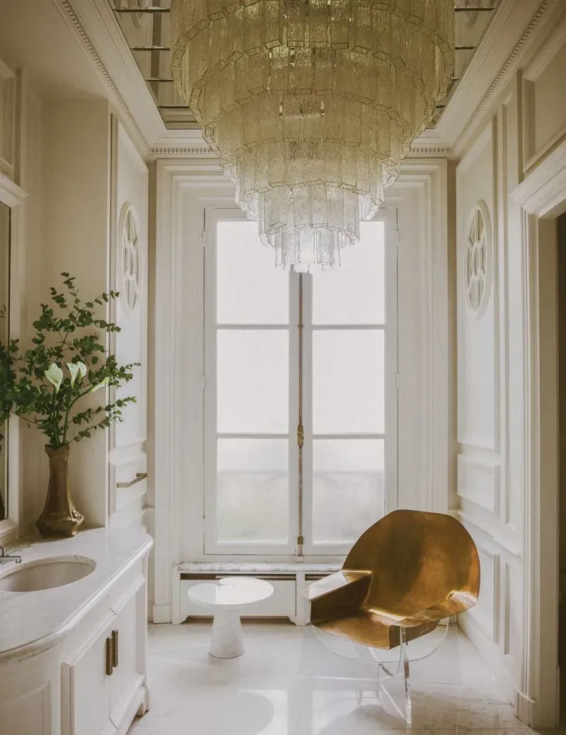 حمام أنيق في منزل ليني كرافيتز في باريس، يحتوي على ثريا زجاجية رائعة، ومغسلة رخامية، وكرسي معدني ذهبي، مع نافذة كبيرة تسمح بدخول الضوء الطبيعي.