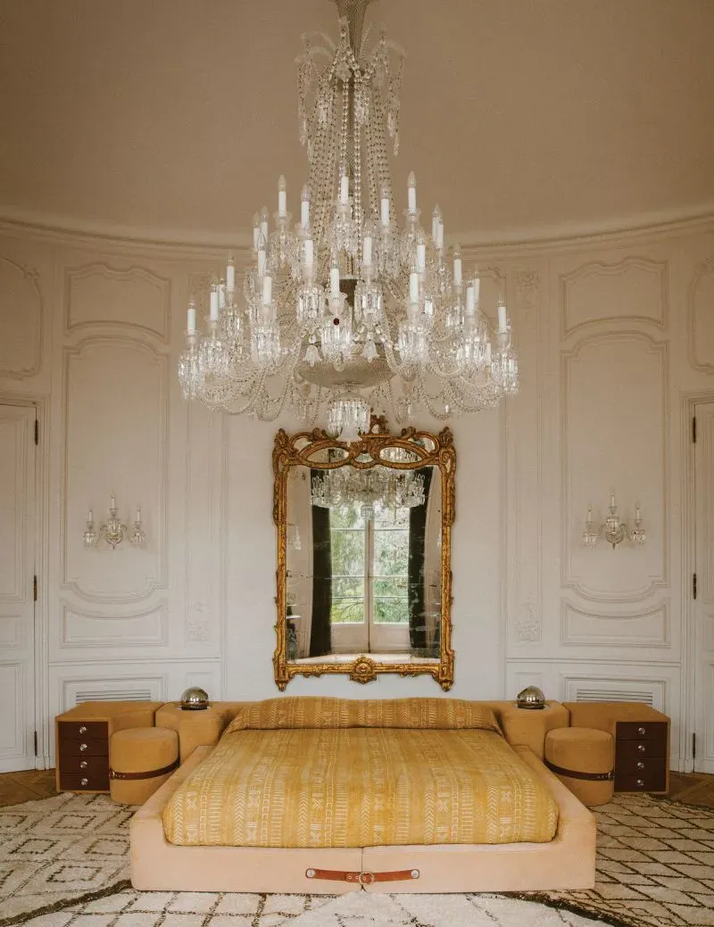 غرفة نوم أنيقة في منزل ليني كرافيتز بباريس، تحتوي على سرير كبير مع غطاء ذهبي، ثريا كريستالية، ومرآة مزخرفة، تعكس أناقته الفريدة.