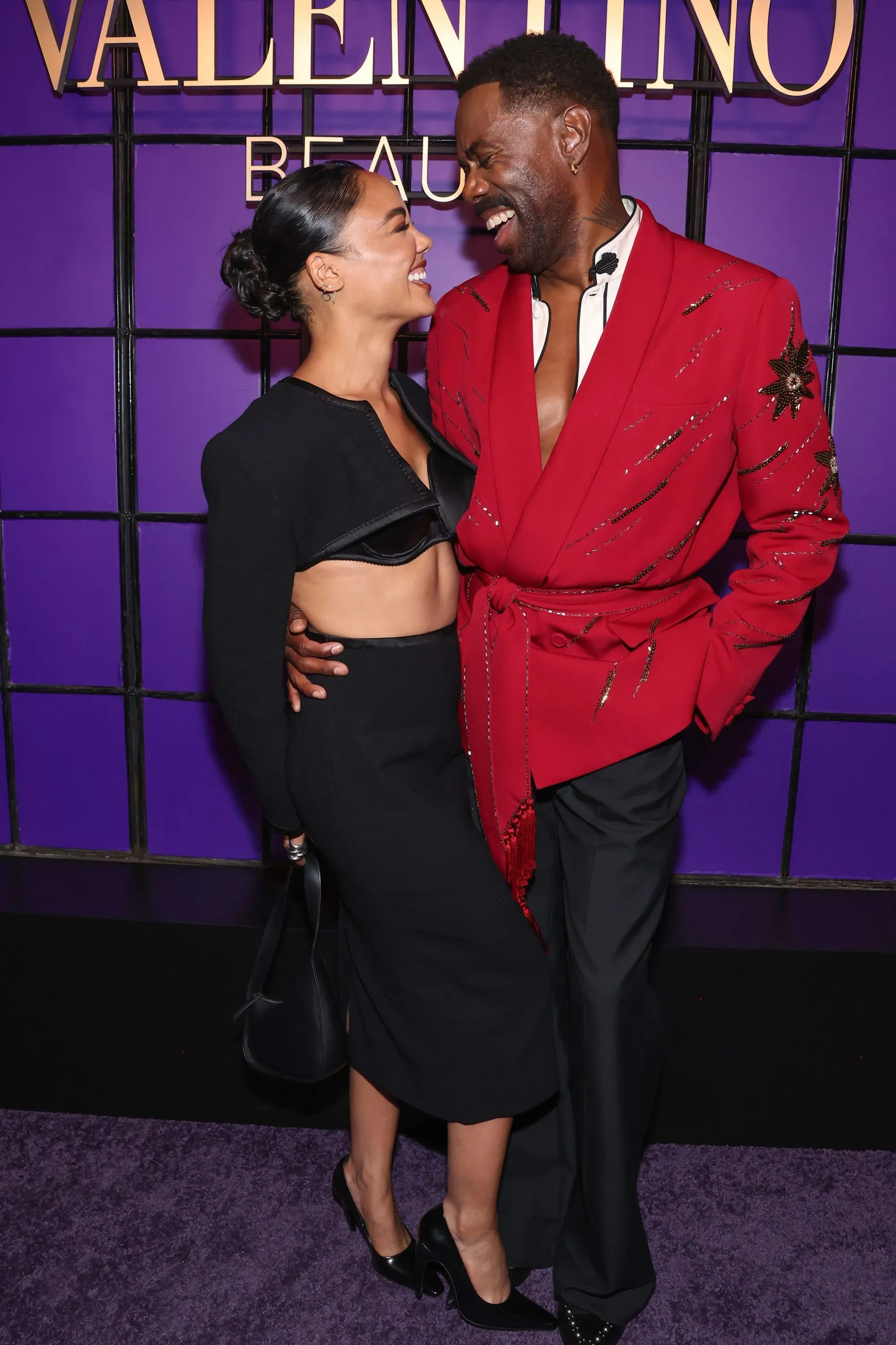 يظهر في الصورة ثنائي مبتسم يتبادلان النظرات في حدث متعلق بماركة فالنتينو، حيث ترتدي المرأة ملابس سوداء بينما الرجل يرتدي سترة حمراء.