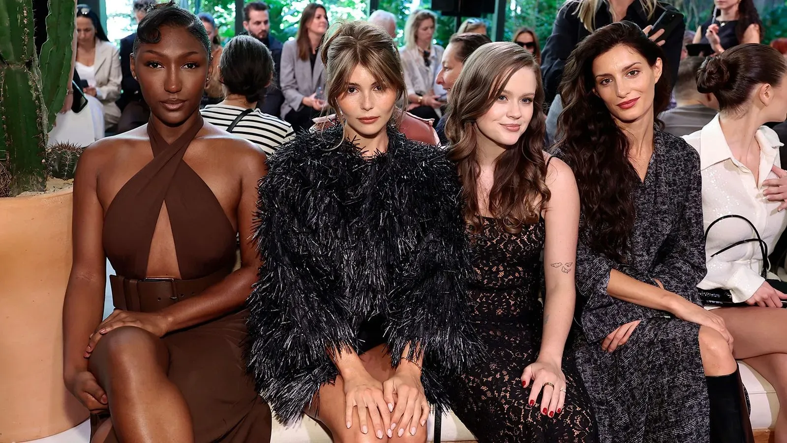 أربع عارضات أزياء يجلسن معاً في عرض أزياء، يرتدين ملابس أنيقة، مع أجواء احتفالية في أسبوع الموضة بنيويورك.