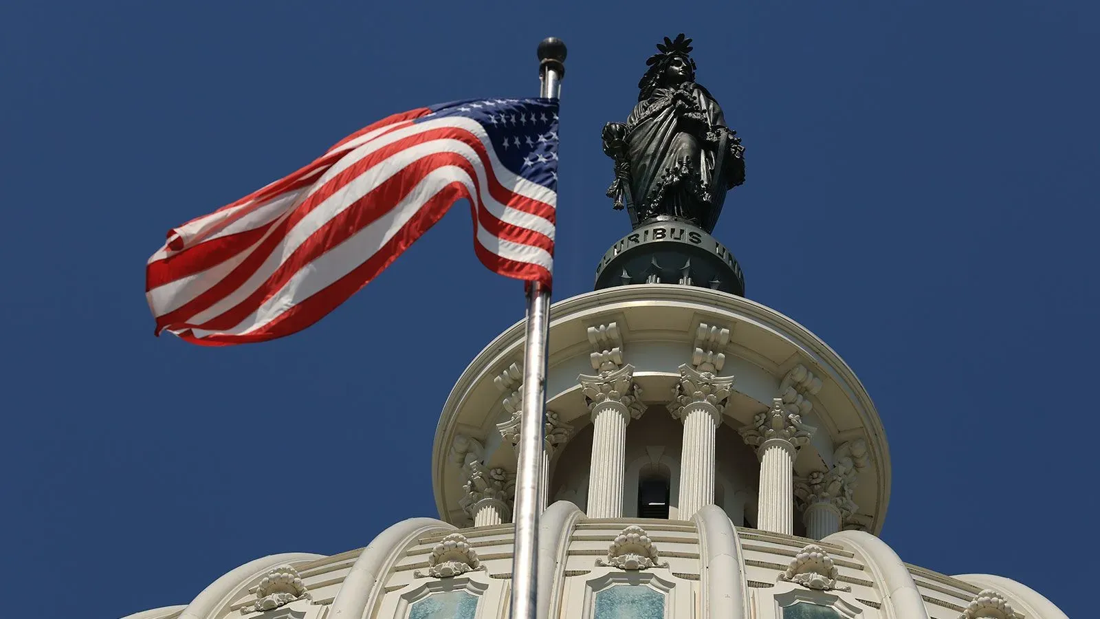 علم الولايات المتحدة يرفرف أمام قبة مبنى الكابيتول، مع تمثال يمثل الحرية في الأعلى، في سماء زرقاء صافية.