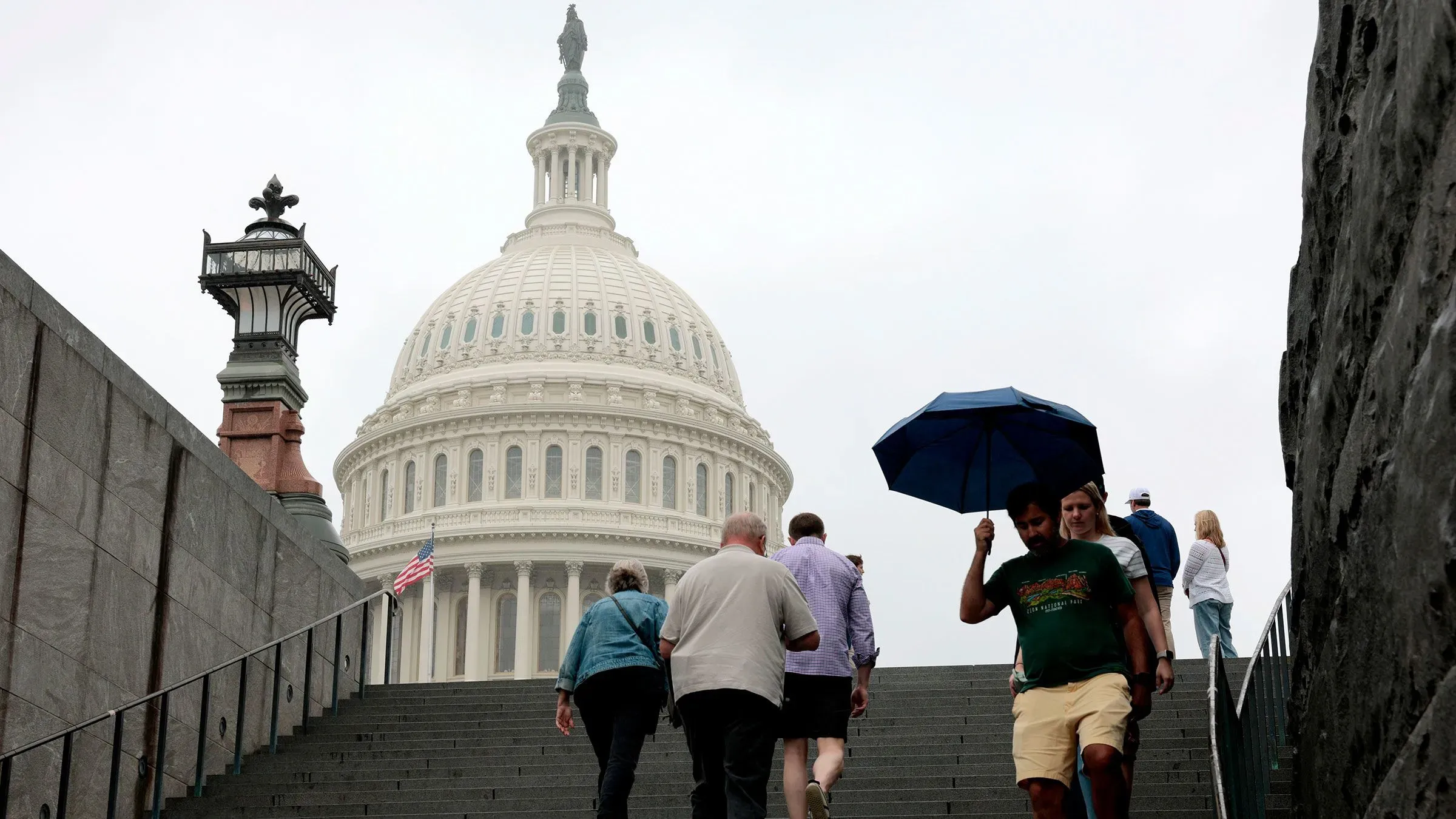 أشخاص يتجهون نحو مبنى الكابيتول الأمريكي تحت سماء غائمة، مع أحدهم يحمل مظلة. يرمز المشهد إلى النشاط السياسي والتشريعي.