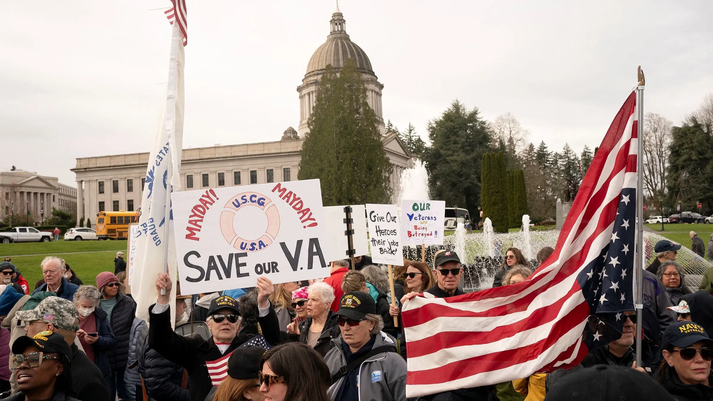 احتجاج أمام مبنى حكومي يطالب بحماية خدمات وزارة شؤون المحاربين القدامى، مع لافتات تعبر عن القلق بشأن التخفيضات المحتملة.