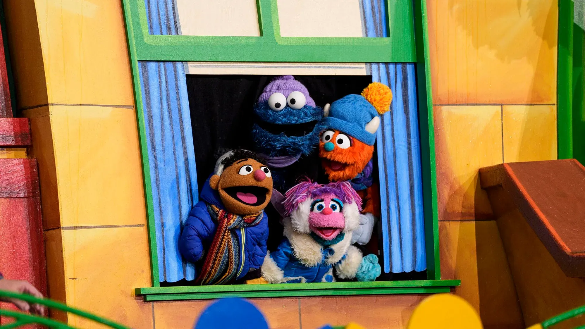 مجموعة من دمى "شارع سمسم" الشهيرة، بما في ذلك إلمو وكوكي مونستر، تتطلع من نافذة ملونة، تعبيراً عن الفرح والترقب لموسمهم الجديد على نتفليكس.