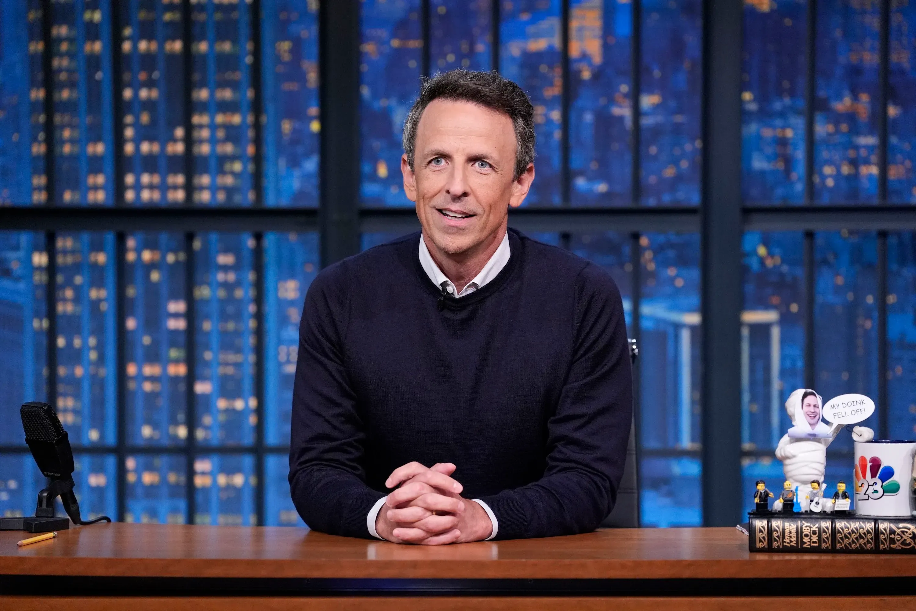 سيث مايرز يجلس خلف مكتبه في برنامجه "آخر الليل"، مع خلفية ليلية من المدينة وديكورات تعكس طابع العرض.