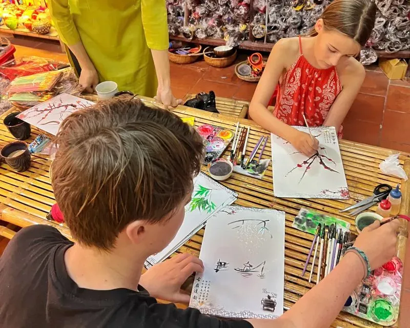 أطفال يتعلمون فن الرسم في ورشة عمل، مع أدوات وأوراق فنية حولهم، مما يعكس تجربة تعليمية ثقافية أثناء السفر.