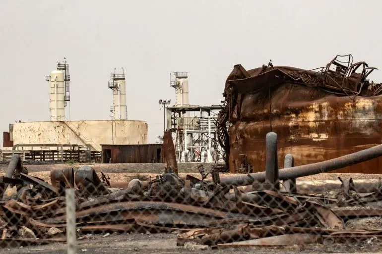 صورة لموقع انفجار في دير الزور، يظهر فيه حطام حافلة متضررة ومنشآت نفطية خلفها، بعد هجوم أسفر عن مقتل أربعة من عناصر الأمن.