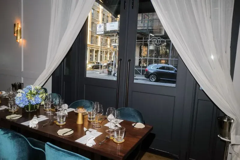 طاولة طعام أنيقة داخل مطعم، مزينة بأزهار زرقاء وأوانٍ زجاجية، مع نوافذ كبيرة تطل على الشارع. تعكس الأجواء الفاخرة للمطعم في مدينة نيويورك.