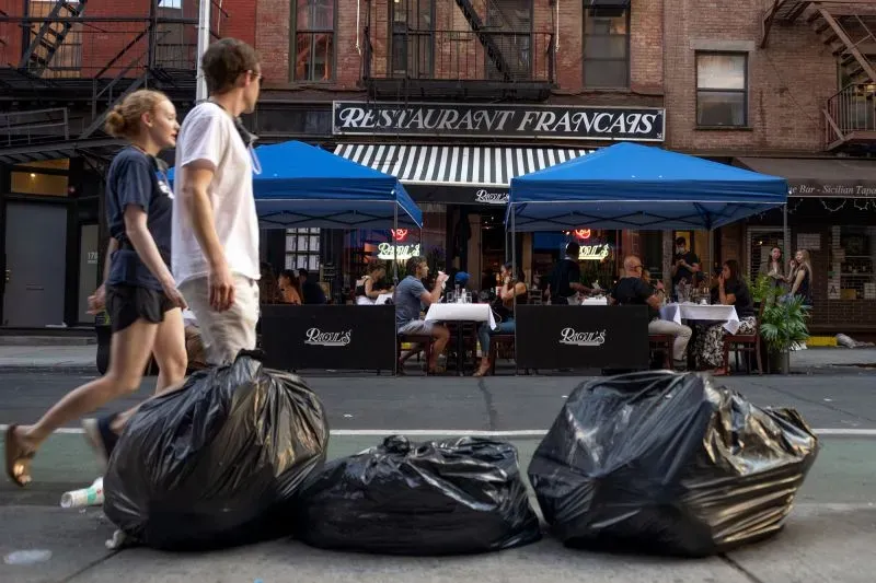 مطعم في نيويورك مع طاولات خارجية تحت خيام زرقاء، بينما يسير الناس بجانب أكياس القمامة، مما يعكس تأثير تناول الطعام في الهواء الطلق.