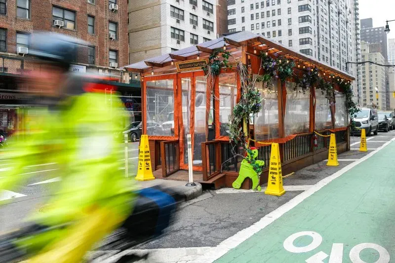 هيكل مطعم خارجي مزين بالأضواء والنباتات، محاط بعلامات تحذيرية، في شارع مزدحم بنيويورك، يعكس تجربة تناول الطعام في الهواء الطلق.