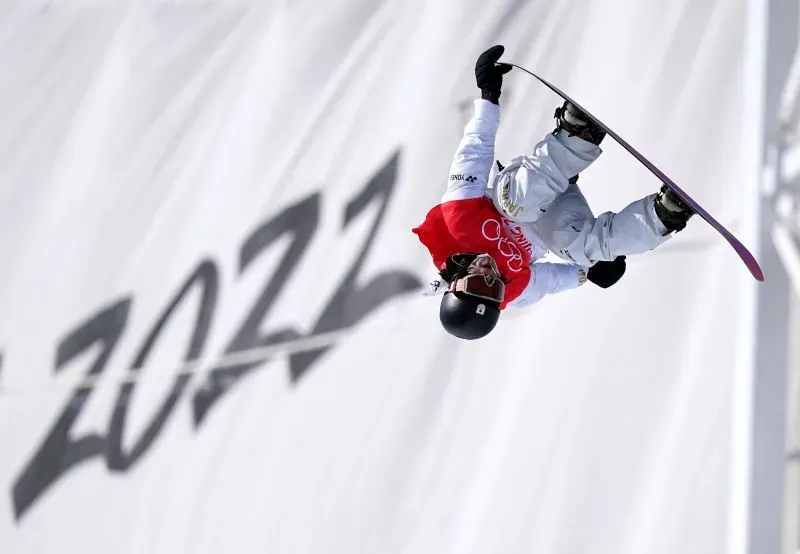 متزلج على الجليد يقوم بحركة بهلوانية في الهواء خلال حدث رياضي، مع وجود شعار الأولمبياد في الخلفية.