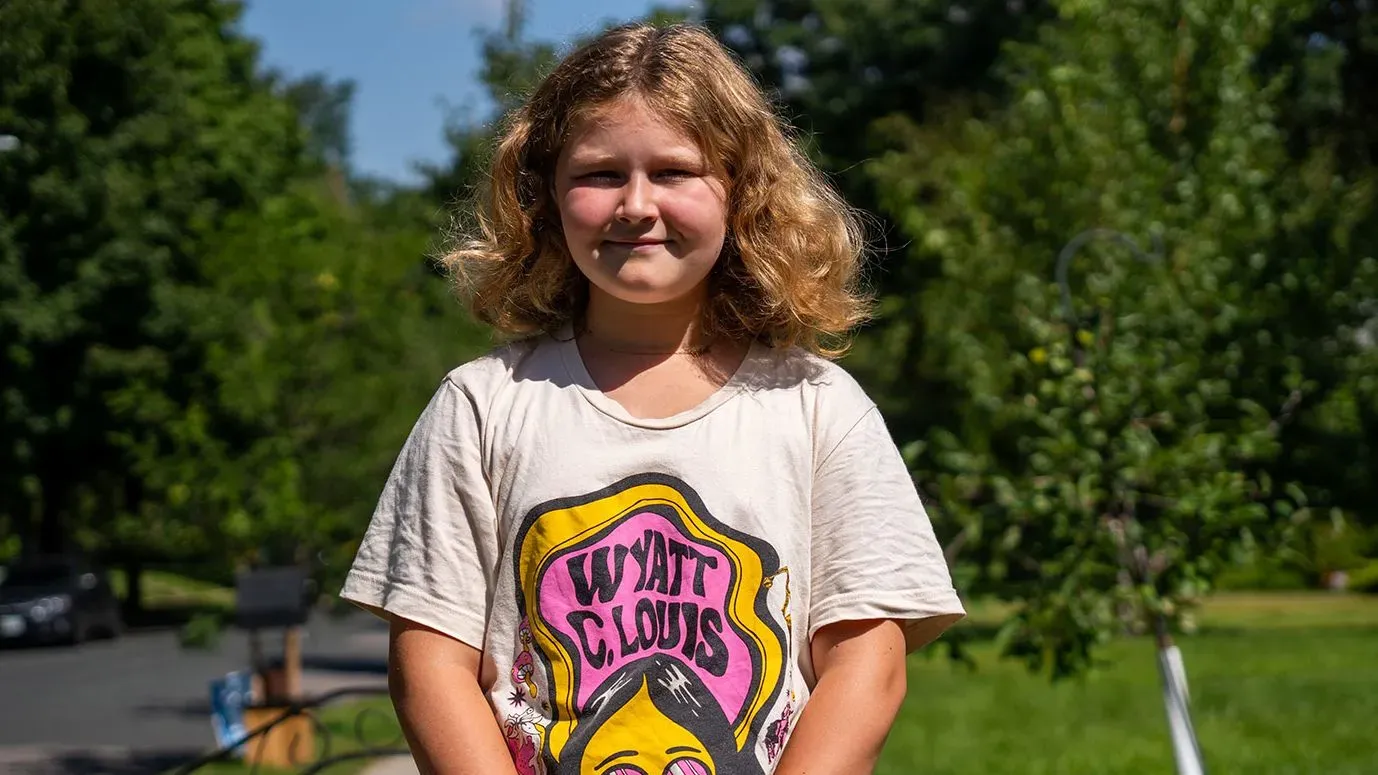 فتاة صغيرة ذات شعر أشقر ومجعد ترتدي قميصًا ملونًا، تقف مبتسمة في حديقة، محاطة بأشجار خضراء. تعكس الصورة براءة الطفولة في ظل أحداث مأساوية.