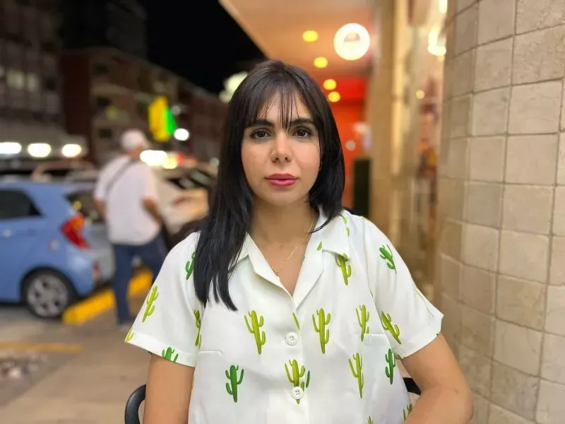 امرأة تجلس في مكان عام، ترتدي قميصًا مزخرفًا بأشكال الصبار، تعكس مشاعر القلق والأمل وسط ظروف الهجرة الصعبة.