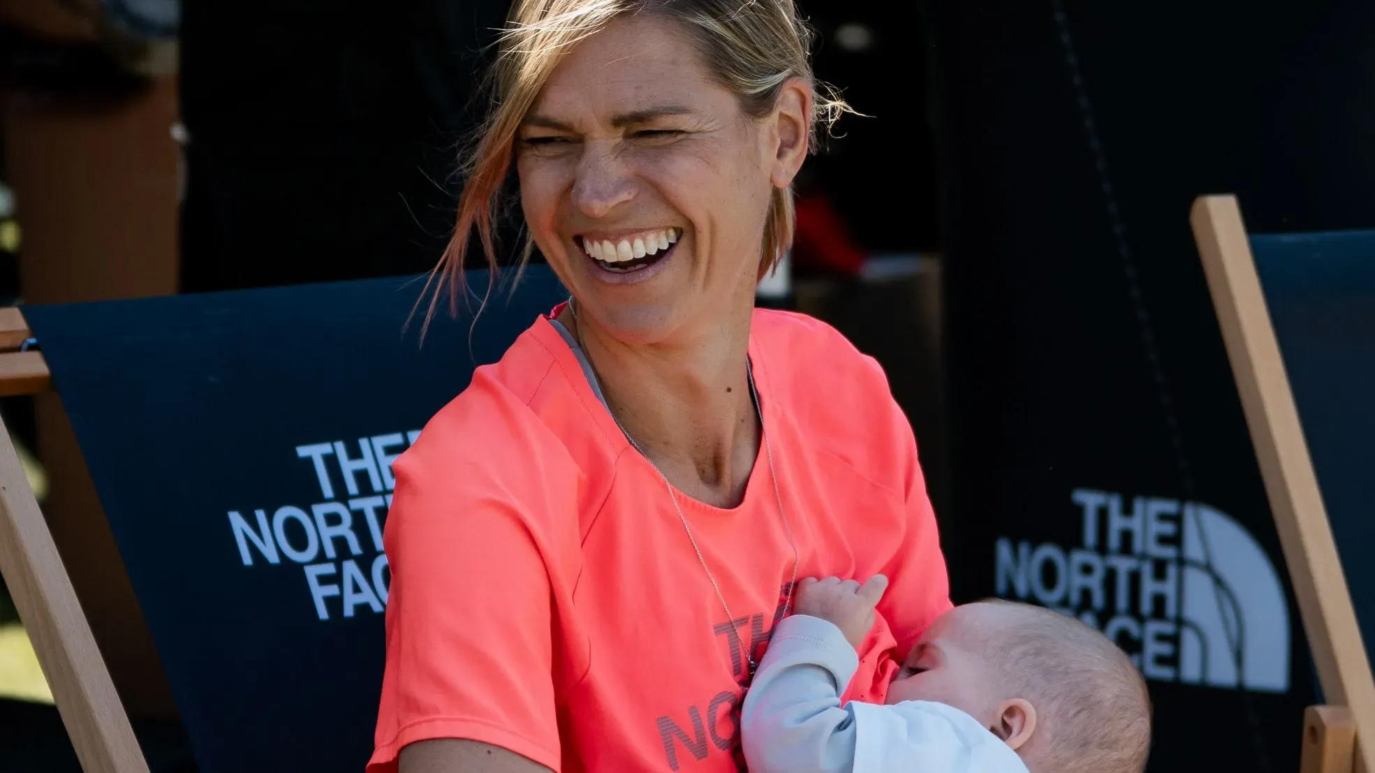 ستيفاني كيس تبتسم بينما ترضع ابنتها بيبر، مرتدية قميص رياضي، في حدث رياضي، مما يعكس التوازن بين الأمومة والرياضة.