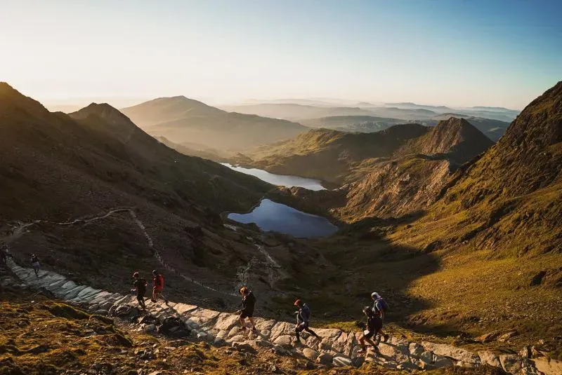 مجموعة من المتسابقين يجتازون مسارًا جبليًا في سباق ألترا تريل سنودونيا بويلز، مع مناظر طبيعية خلابة للجبال والبحيرات.