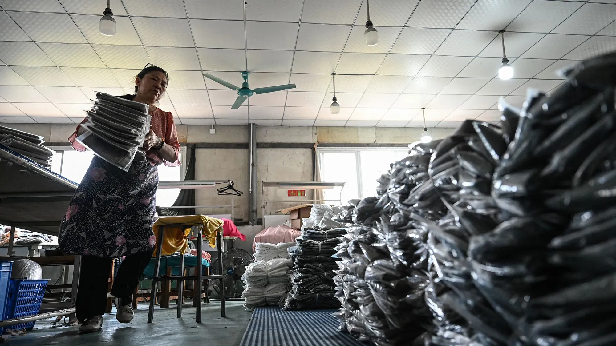 امرأة تعمل في مصنع، تحمل أكوامًا من الملابس المعبأة، مع وجود أكوام إضافية من الملابس على الأرض.