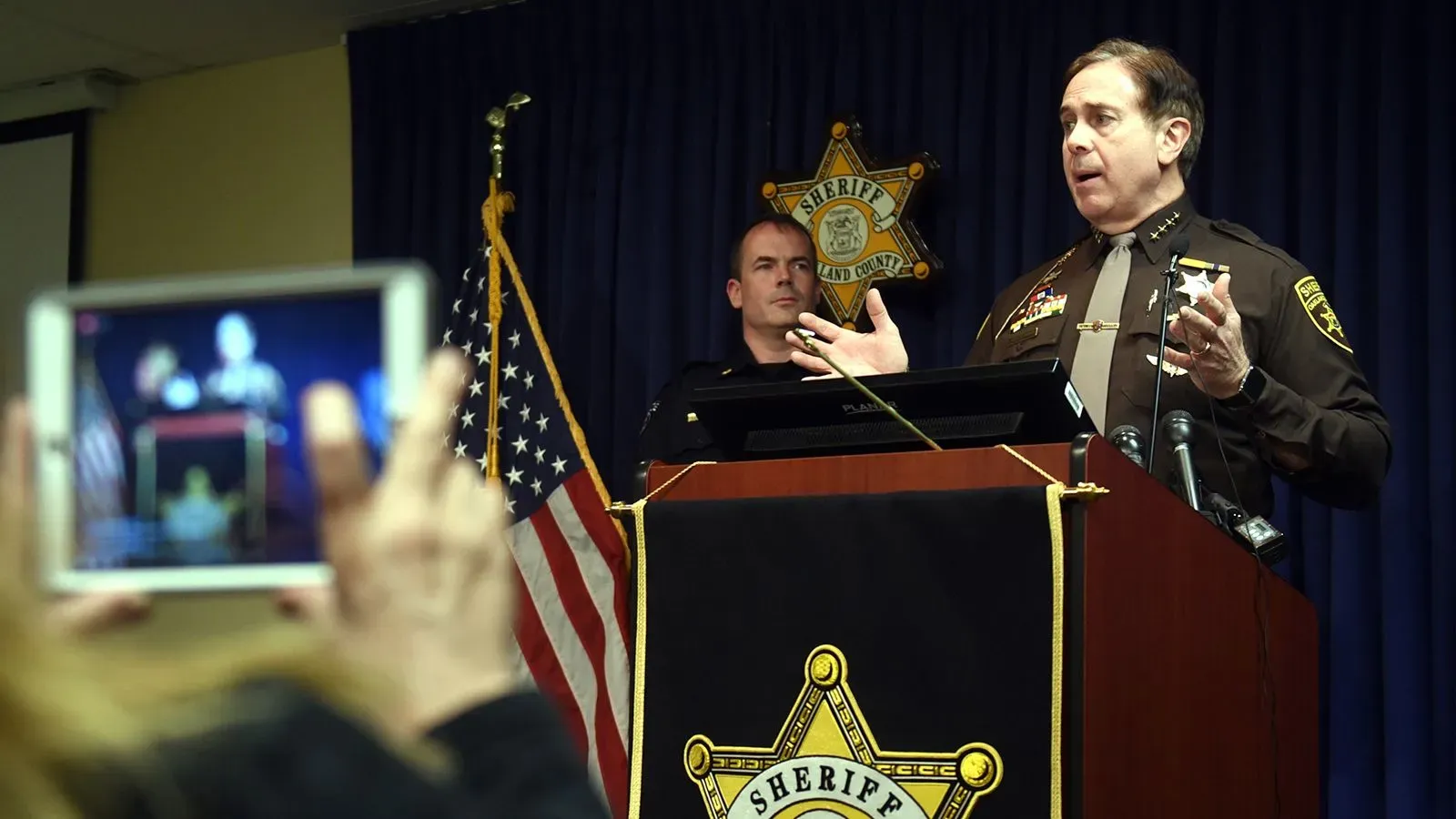 عمدة مقاطعة أوكلاند يتحدث في مؤتمر صحفي حول حالة الأطفال المهجورين، مع وجود ضابط شرطة خلفه.