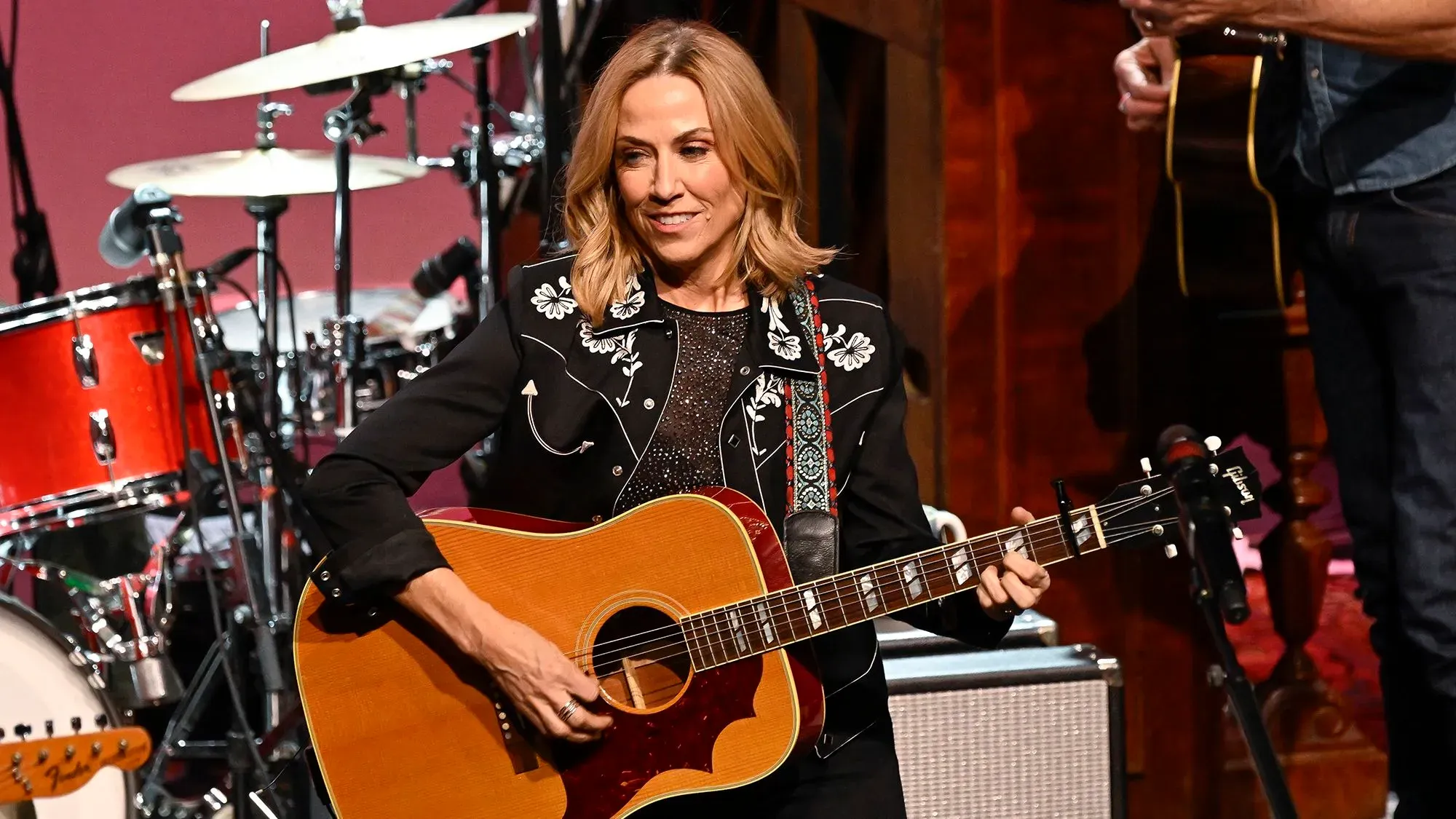 شيريل كرو تعزف على الغيتار خلال عرض موسيقي، مع خلفية من الطبول، تعبيرًا عن احتجاجها على إيلون ماسك.