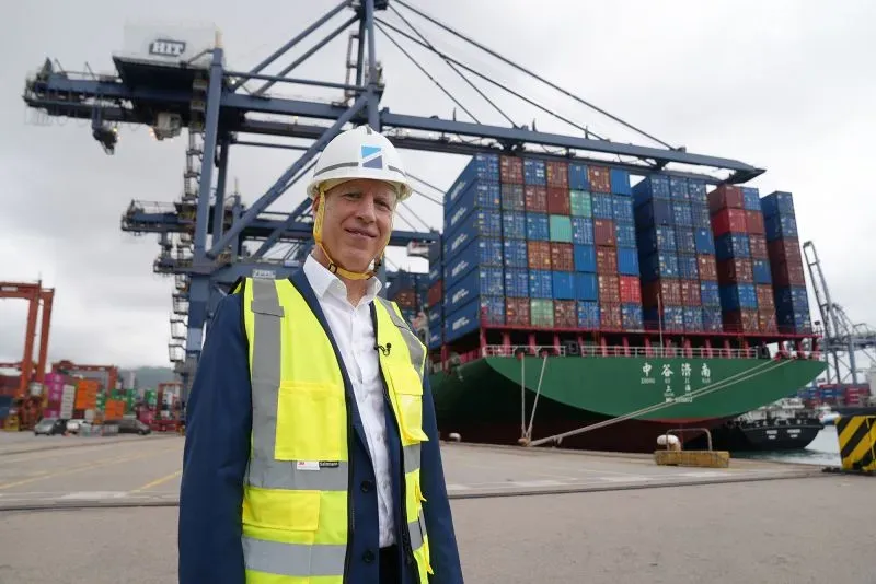 رجل يرتدي سترة واقية وخوذة، يقف أمام سفينة حاويات ضخمة محملة بأكثر من 700 حاوية، في ميناء هونغ كونغ.