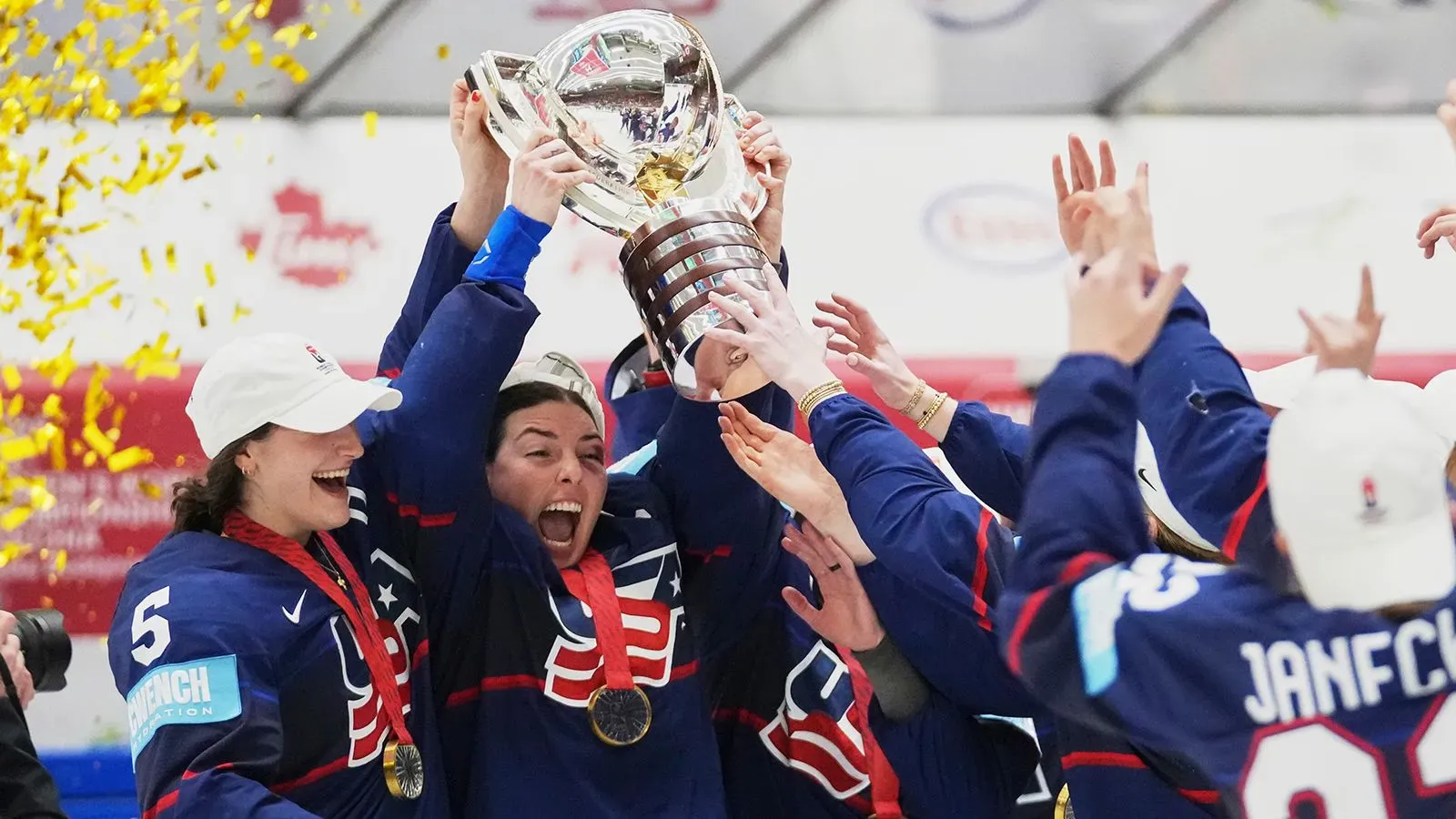 فريق الولايات المتحدة يحتفل بفوزهم ببطولة العالم لهوكي الجليد للسيدات، رافعين الكأس مع تساقط قصاصات الذهب في الخلفية.