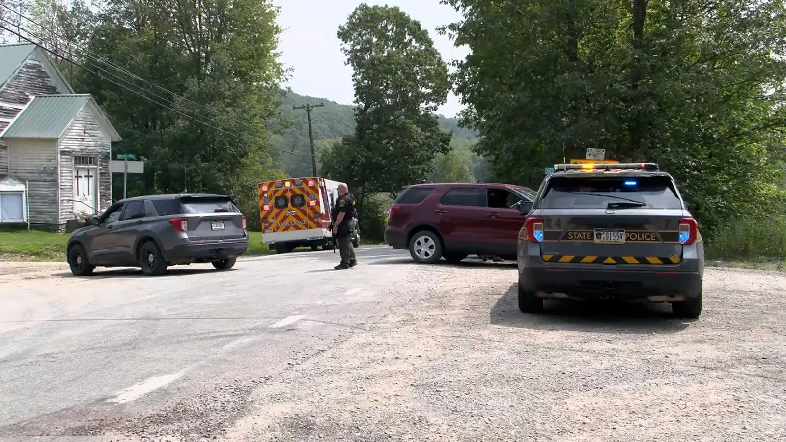 استجابة الطوارئ في مكان حادث إطلاق نار، تظهر سيارات الشرطة والإسعاف في منطقة ريفية، مع وجود شرطي يتفقد الموقع.