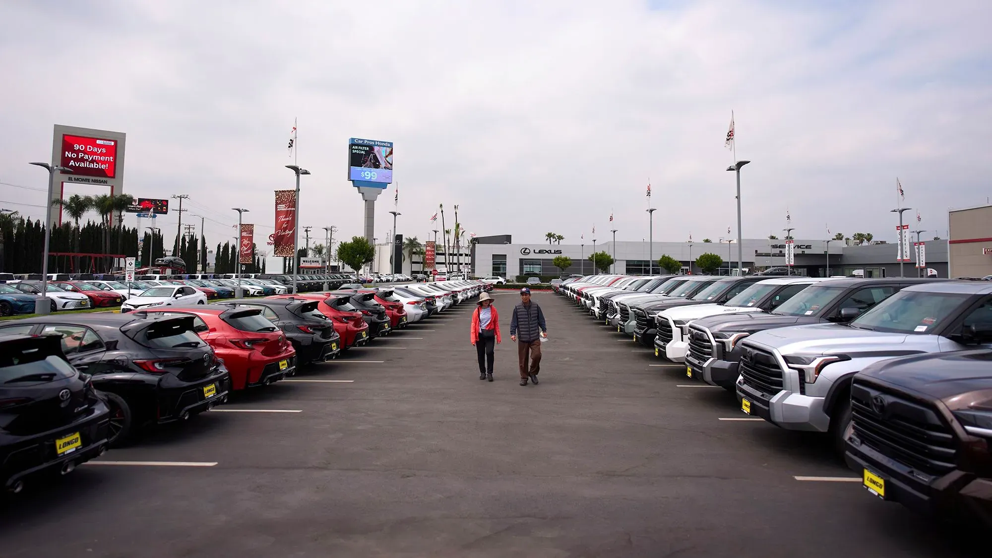 ازدحام في ساحة عرض سيارات، حيث يتجول شخصان بين صفوف من السيارات الجديدة، مع لافتات ترويجية في الخلفية تعلن عن عروض خاصة.