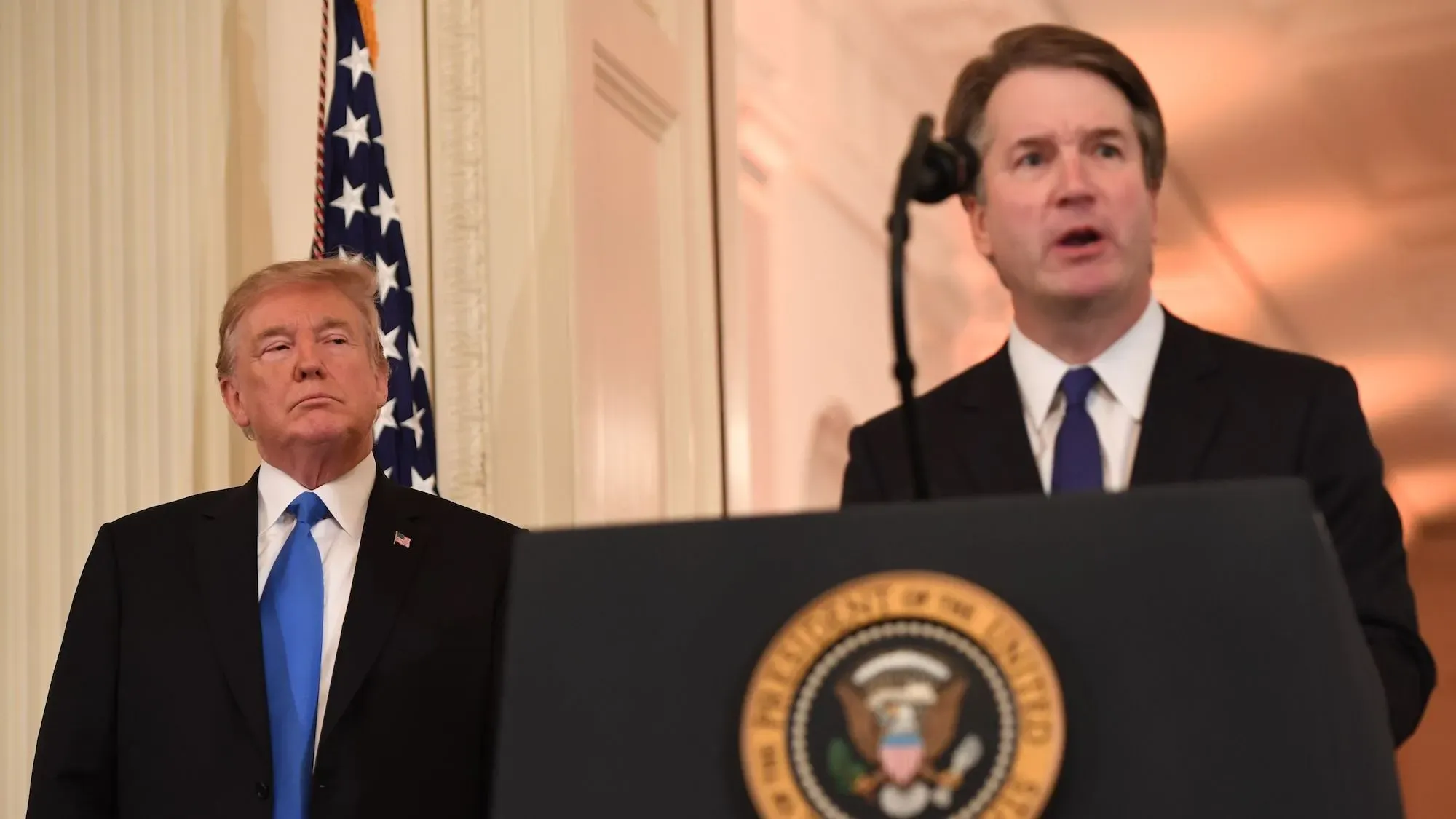 دونالد ترامب يقف خلف ميكروفون أثناء خطاب القاضي بريت كافانو في حفل التنصيب، مع العلم الأمريكي خلفهم، مشيراً إلى التحولات السياسية في المحكمة العليا.