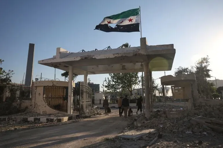 مدخل مدمّر لموقع عسكري في سوريا، يرفرف فوقه علم الثورة السورية، مع أشخاص يسيرون في الخلفية.
