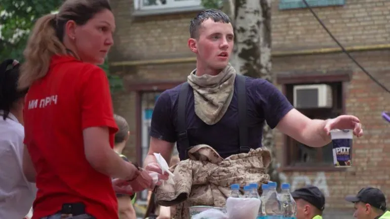 شخصان يعملان معًا في موقع دمار، حيث يقدم أحدهما المساعدة بينما يحمل الآخر كوبًا. تعكس الصورة جهود الإغاثة في أوكرانيا.