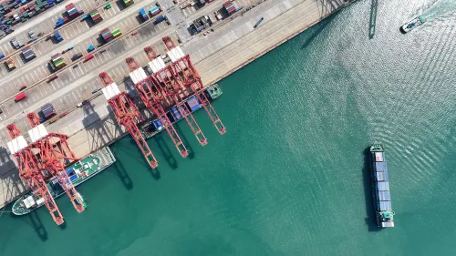 صورة جوية لميناء يحتوي على رافعات حمراء وحاويات شحن، مع سفينة تبحر في المياه القريبة، تعكس حركة التجارة الدولية.