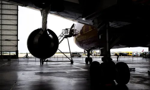 صورة لمطار يظهر فيها طائرة تحت الصيانة مع مدرج خلفي، حيث يعمل عمال في بيئة صناعية. تتعلق الصورة بتحقيقات حول تفجيرات طرود في أوروبا.