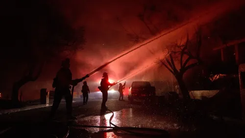 رجال الإطفاء يعملون على إخماد حريق في منطقة سكنية، مع أضواء الطوارئ في الخلفية والدخان الكثيف يملأ السماء، مما يعكس تأثير أزمة المناخ.