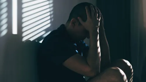 رجل يجلس في مكان مظلم، ممسكًا برأسه بيديه، مع تسليط الضوء على مشاعره من خلال الظلال، مما يعكس الصراع الداخلي والمعاناة.
