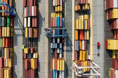 صورة جوية لميناء يحتوي على حاويات شحن ملونة مرتبة بشكل منظم، مع شاحنة صغيرة تسير بين الحاويات، تعكس حركة التجارة العالمية.