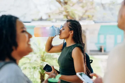 امرأة تشرب من زجاجة مياه قابلة لإعادة الاستخدام في حرم جامعي، محاطة بأصدقاء، مما يعكس أسلوب حياة مستدام واهتمامًا بالبيئة.