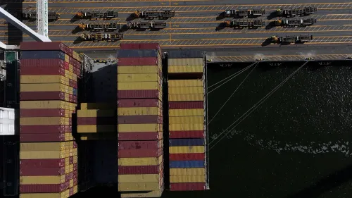 صورة توضح حاويات شحن ملونة في ميناء، مع شاحنات مخصصة لنقل البضائع، تعكس تأثير الحرب التجارية على الاقتصاد العالمي.