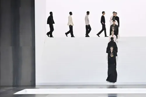 عرض أزياء رجالية في باريس يظهر مجموعة من العارضين يسيرون على منصة بيضاء، مع تركيز على الأزياء الأنيقة والتصاميم الحديثة.