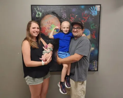 عائلة فيلا تتوسط الصورة، حيث يحمل الأب كودي ابنه كايلين المصاب بالسرطان، بينما تحمل الأم آلي ابنتهم حديثة الولادة، مع خلفية فنية ملونة.