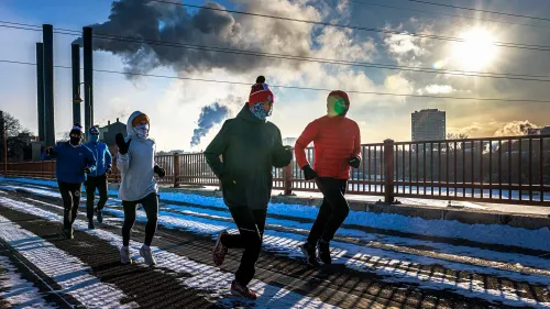 مجموعة من العدائين يرتدون ملابس شتوية، يجري في منطقة مغطاة بالثلوج، مع ظهور مدخنة تدخين في الخلفية وأشعة الشمس الساطعة.