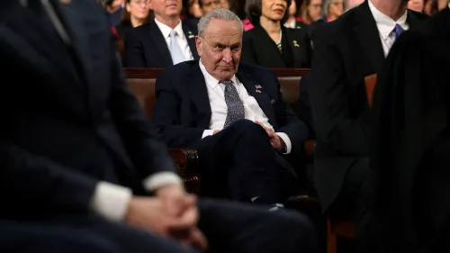 تشاك شومر، زعيم الديمقراطيين في مجلس الشيوخ، يجلس بتعبير جاد خلال حدث رسمي، مع أعضاء آخرين في الخلفية، مما يعكس ضغوط السياسة الحالية.