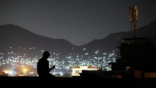 شخص يجلس في الظلام على سطح، مستخدمًا هاتفه، بينما تظهر أضواء المدينة خلفه، مما يبرز تأثير انقطاع الإنترنت في أفغانستان.