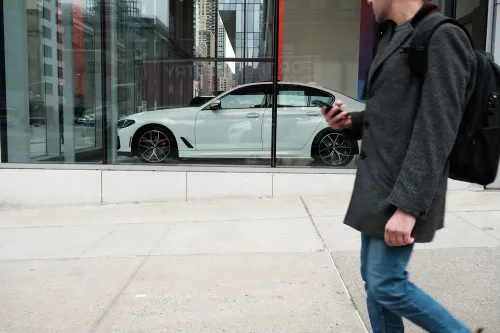 رجل يمشي بجانب واجهة عرض تحتوي على سيارة بيضاء حديثة، تعكس الضغوط المالية وارتفاع تكاليف المعيشة في الولايات المتحدة.