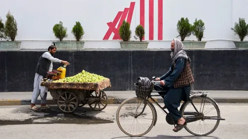 رجل يبيع التفاح على عربة في الشارع، بينما يمر رجل آخر على دراجة هوائية، مع خلفية جدارية تحمل شعاراً.