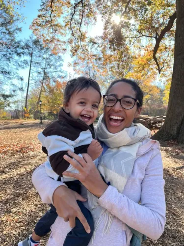 امرأة تحمل طفلها في حضنها، مبتسمة في حديقة مليئة بالأشجار الملونة، تعكس لحظات من السعادة والتواصل الأسري.