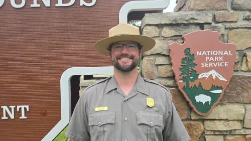 براين غيبس يرتدي زي حارس الحديقة أمام لافتة النصب التذكاري الوطني لتلال إفيجي، حيث كان يعمل على تعليم الزوار عن تاريخ الموقع.